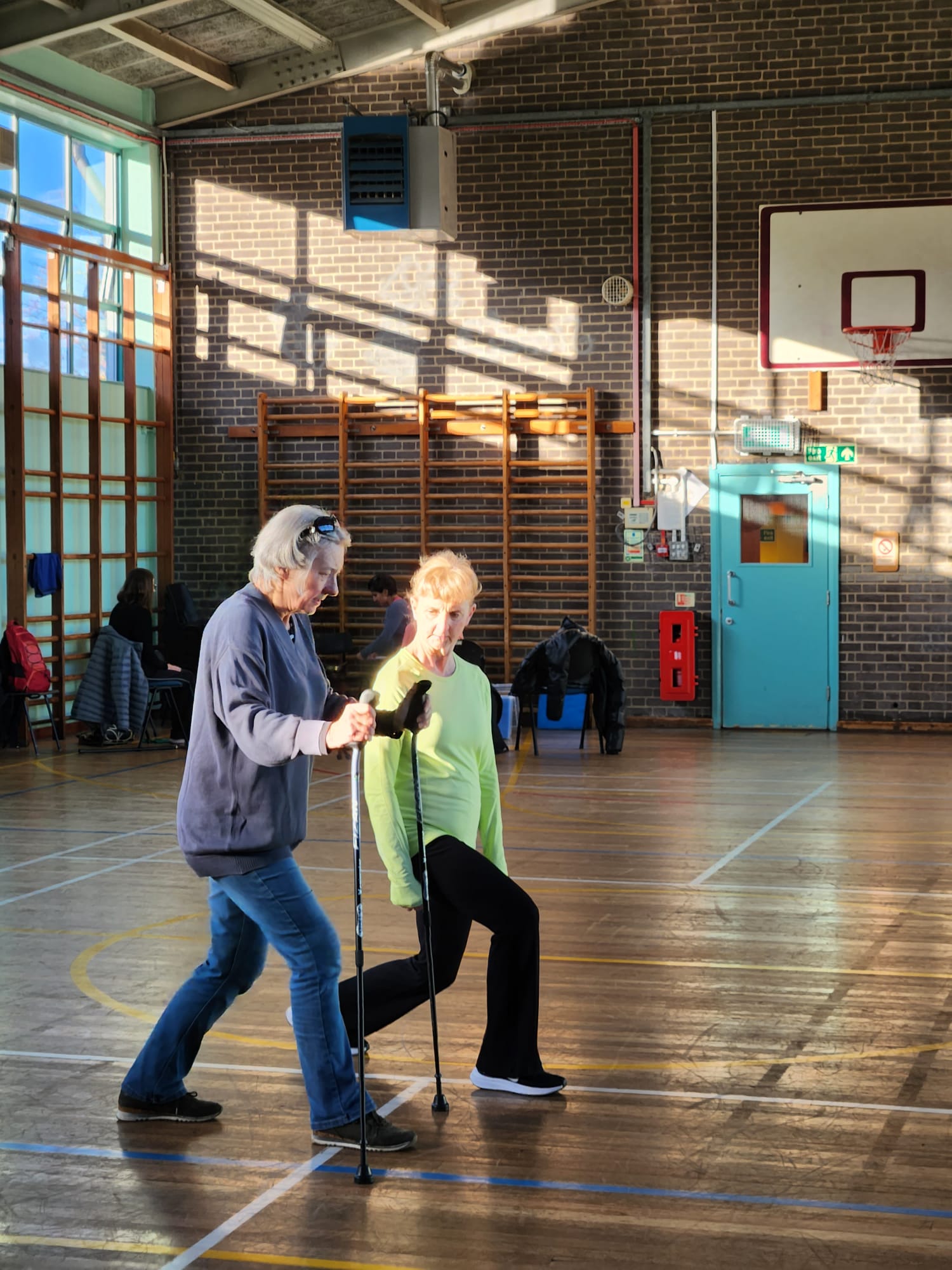 Two people walking in a gymnasium with wooden floors and brick walls.